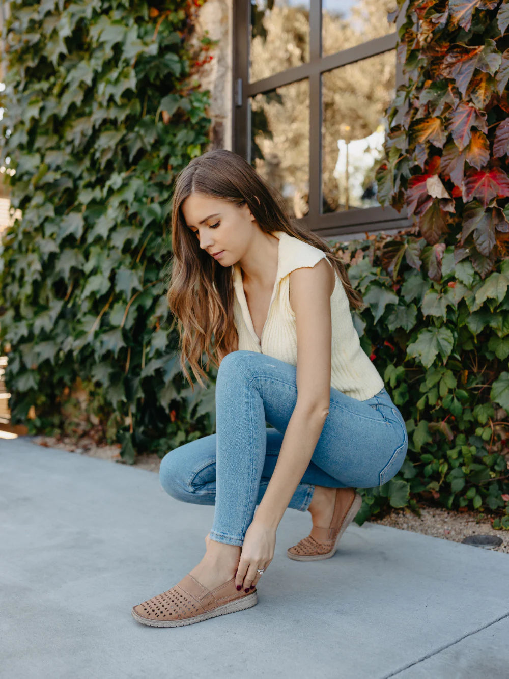 Woman in light-colored top and blue jeans sitting on a concrete surface with greenery in the background